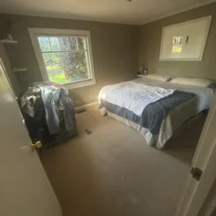 Before photo of a residential bedroom flooring project by Interstate Flooring on SW Pomona St in Portland, Oregon. The image shows outdated beige carpeting, olive-green walls, a large bed, and a window overlooking the yard before a carpet replacement or flooring upgrade