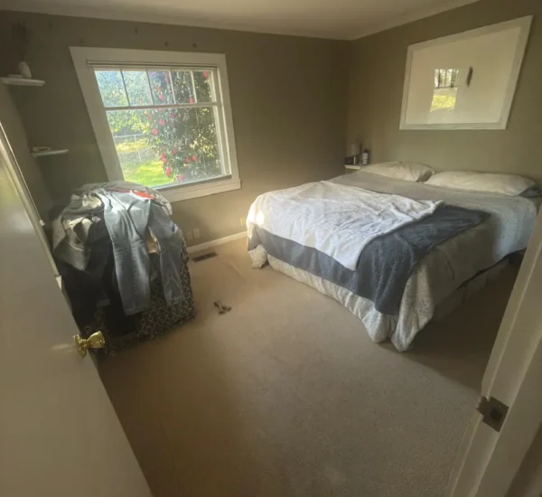 Before photo of a residential bedroom flooring project by Interstate Flooring on SW Pomona St in Portland, Oregon. The image shows outdated beige carpeting, olive-green walls, a large bed, and a window overlooking the yard before a carpet replacement or flooring upgrade
