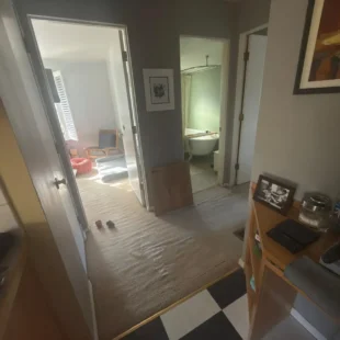 Before photo of a home flooring upgrade by Interstate Flooring on SW Pomona St in Portland, Oregon. Shows loose, heavily rippled beige hallway carpet transitioning to a checkered kitchen floor, with a wood flooring sample board leaning against the wall before carpet removal