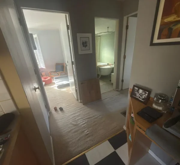 Before photo of a home flooring upgrade by Interstate Flooring on SW Pomona St in Portland, Oregon. Shows loose, heavily rippled beige hallway carpet transitioning to a checkered kitchen floor, with a wood flooring sample board leaning against the wall before carpet removal