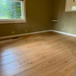 After photo of a residential bedroom flooring remodel by Interstate Flooring on SW Pomona St in Portland, Oregon. Shows newly installed wide-plank engineered hardwood flooring in a light oak finish, contrasting with olive-green walls and clean white trim