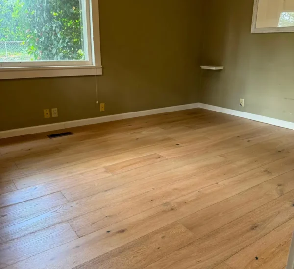 After photo of a residential bedroom flooring remodel by Interstate Flooring on SW Pomona St in Portland, Oregon. Shows newly installed wide-plank engineered hardwood flooring in a light oak finish, contrasting with olive-green walls and clean white trim