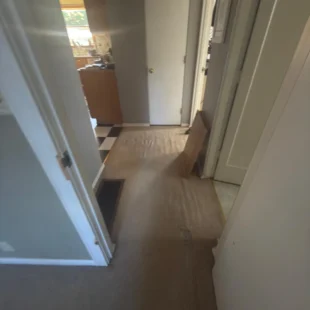 Before photo of a residential hallway flooring renovation by Interstate Flooring on SW Pomona St in Portland, Oregon. Shows worn, rippled beige carpeting in a narrow corridor leading to a kitchen with black-and-white checkered flooring, prior to a hardwood or luxury vinyl flooring upgrade
