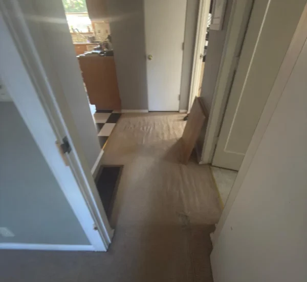 Before photo of a residential hallway flooring renovation by Interstate Flooring on SW Pomona St in Portland, Oregon. Shows worn, rippled beige carpeting in a narrow corridor leading to a kitchen with black-and-white checkered flooring, prior to a hardwood or luxury vinyl flooring upgrade