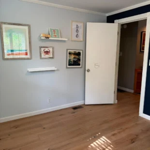 After photo of a bedroom flooring renovation by Interstate Flooring on SW Pomona St in Portland, Oregon. Shows newly installed light oak hardwood floors, light gray walls decorated with framed coastal artwork, and an open white door showing the transition to the hallway