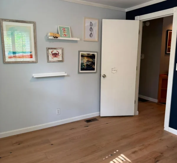 After photo of a bedroom flooring renovation by Interstate Flooring on SW Pomona St in Portland, Oregon. Shows newly installed light oak hardwood floors, light gray walls decorated with framed coastal artwork, and an open white door showing the transition to the hallway