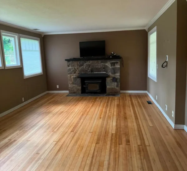 Completed hardwood flooring installation by Interstate Flooring at the SW Pomona St project in Portland, Oregon. Shows a spacious living room featuring newly polished oak hardwood floors, taupe walls with white trim, and a rustic stone fireplace