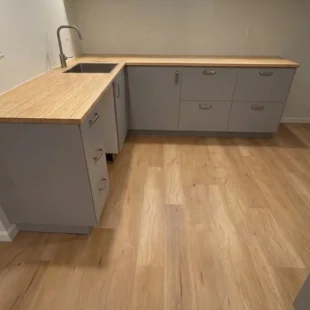 Newly installed wood-look luxury vinyl plank flooring in a modern finished basement kitchenette by Interstate Flooring on N Halleck St in Portland, featuring light grey lower cabinets, butcher block countertops, and a undermount sink