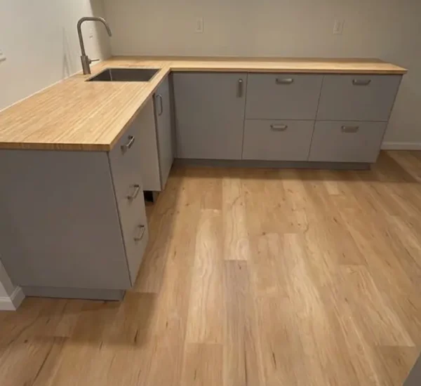 basement-flooring-installation-kitchenette-n-halleck-portland Newly installed wood-look luxury vinyl plank flooring in a modern finished basement kitchenette by Interstate Flooring on N Halleck St in Portland, featuring light grey lower cabinets, butcher block countertops, and a undermount sink