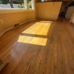 Before photo of a dining area hardwood floor refinishing project on SW Ridgewood Ave in Portland, showing dull, discolored oak wood flooring with surface scratches under bright window sunlight next to a kitchen island