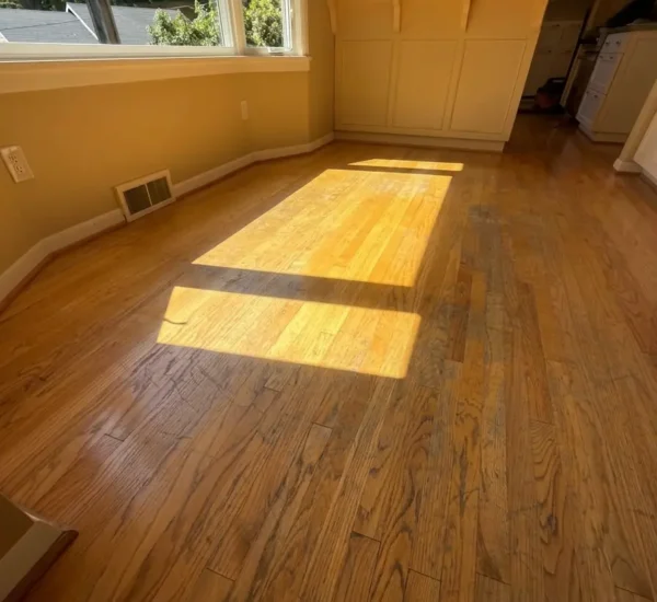 hardwood-floor-refinishing-dining-area-before-sw-ridgewood-ave-portland Before photo of a dining area hardwood floor refinishing project on SW Ridgewood Ave in Portland, showing dull, discolored oak wood flooring with surface scratches under bright window sunlight next to a kitchen island