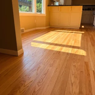 Refinished natural oak hardwood flooring in a sunlit dining area adjacent to a kitchen island by Interstate Flooring on SW Ridgewood Ave in Portland, showing bright sunlight streaming through large windows onto clear-finished wood planks.
