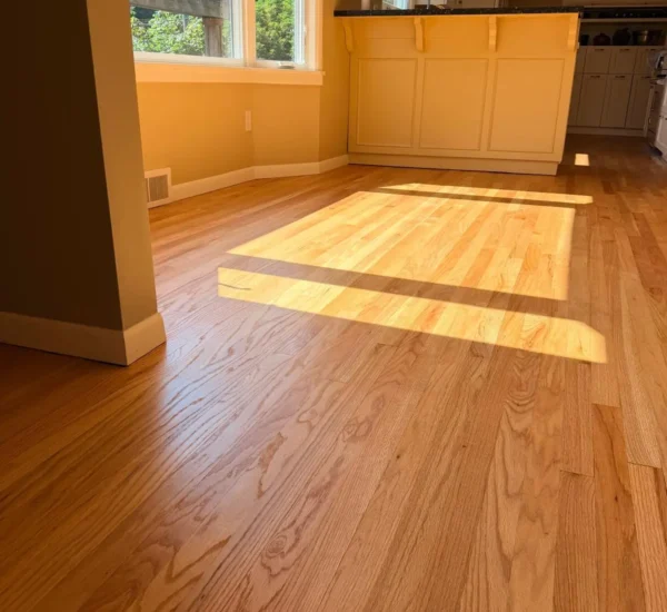 hardwood-floor-refinishing-dining-area-sw-ridgewood-ave-portland Refinished natural oak hardwood flooring in a sunlit dining area adjacent to a kitchen island by Interstate Flooring on SW Ridgewood Ave in Portland, showing bright sunlight streaming through large windows onto clear-finished wood planks.