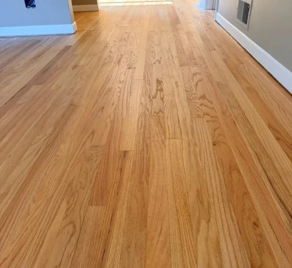 hardwood-floor-refinishing-hallway-sw-ridgewood-ave-portland Refinished red oak hardwood flooring in a residential hallway leading toward a bright kitchen by Interstate Flooring on SW Ridgewood Ave in Portland, showcasing fine wood grain detail, smooth satin finish, and clean white baseboards.