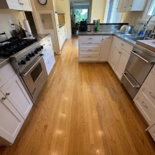 Before photo of a kitchen aisle hardwood floor refinishing project on SW Ridgewood Ave in Portland, showing aged oak wood floors with a dull yellowed finish between white cabinetry and stainless steel appliances