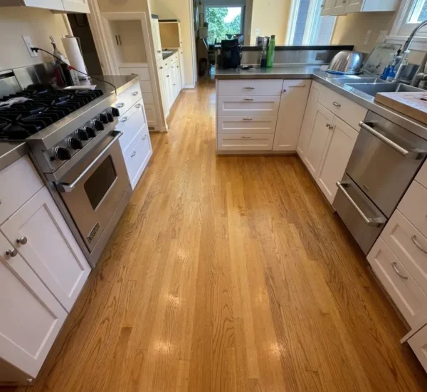 hardwood-floor-refinishing-kitchen-aisle-before-sw-ridgewood-ave-portland Before photo of a kitchen aisle hardwood floor refinishing project on SW Ridgewood Ave in Portland, showing aged oak wood floors with a dull yellowed finish between white cabinetry and stainless steel appliances