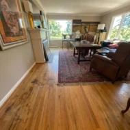 hardwood-floor-refinishing-living-room-before-sw-ridgewood-ave-portland Before photo of a living room hardwood floor refinishing project on SW Ridgewood Ave in Portland, showing heavily worn oak wood flooring with dark water stains and scratches next to a fireplace and area rug