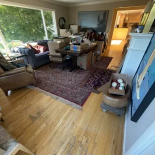 Before photo of a living room hardwood floor refinishing project on SW Ridgewood Ave in Portland, showing packed boxes, furniture prep, and heavily worn oak floors with finish loss and dark scuff marks