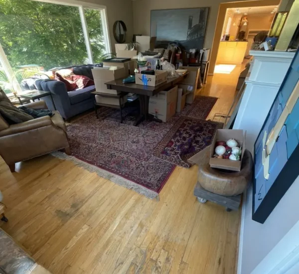 hardwood-floor-refinishing-living-room-prep-before-sw-ridgewood-ave-portland Before photo of a living room hardwood floor refinishing project on SW Ridgewood Ave in Portland, showing packed boxes, furniture prep, and heavily worn oak floors with finish loss and dark scuff marks