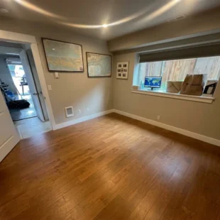 hardwood-flooring-home-office-portland-or Finished home office space featuring warm oak hardwood flooring, white baseboard trim, framed wall art, and a large daylight window in Portland, Oregon